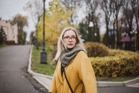 Portrait of a young woman in glasses with long blond hair in a yellow coat in an autumn parkの写真素材