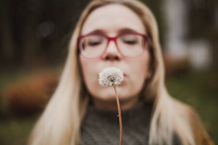 Cute blonde in glasses blowing on dandelions in autumn parkの写真素材