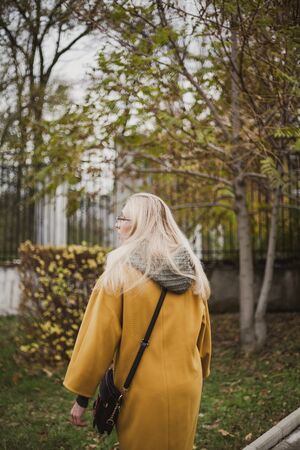 slender blonde in a bright yellow fashion coat walks in an autumn park. View from the backの写真素材