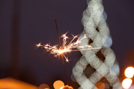 Sparklers burn in male hands against backdrop of burning urban Christmas tree.の写真素材