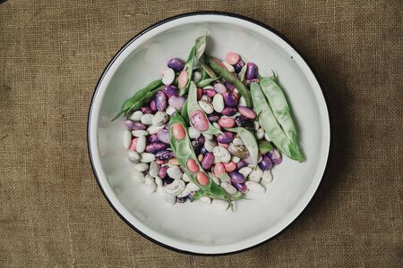 Pods and beans of different colors lie in a white round metal bowl. View from above.の写真素材