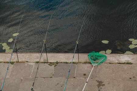 Fishing in the city. Three long fishing rods are installed on the concrete embankment on the shore of the reservoir. There is a green net nextの写真素材