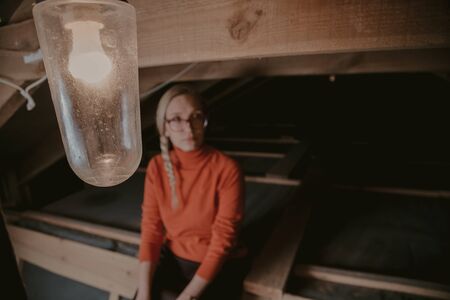 Pretty blonde in an orange sweater sits on wooden boards in the attic. Raised legs high in leather bootsの写真素材