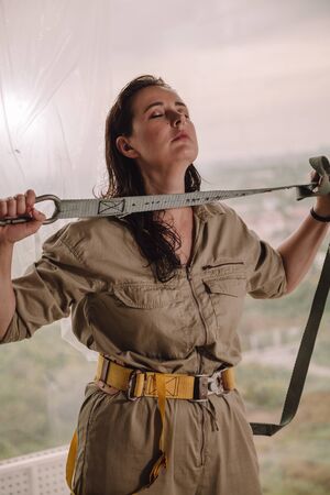 Nice girl in gray overalls holds in her hands a safety belt with a carbine on the background of the window. Outside the window a beautiful view and a white spotの写真素材