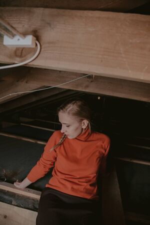 pretty blonde in glasses and an orange sweater sits on wooden boards in the attic