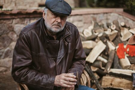 Portrait of a respectable man. An elderly man with a beard in a leather jacket and cap holds a glass...