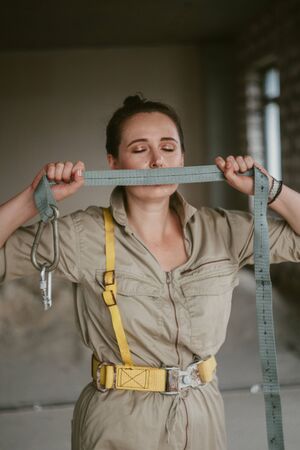 Silent beauty. Nice girl in gray overalls closes her mouth with a safety belt with a carbine on the background of the window. Female portrait without retouching with her natural imperfections.の写真素材