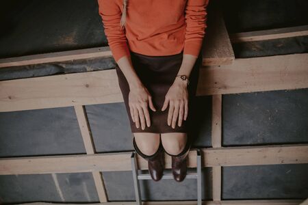 Pretty blonde in an orange sweater and leather boots sits on a structure from wooden planks in the atticの写真素材