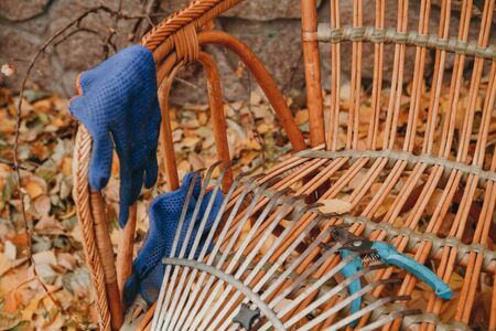 Autumn still life. A fan rake and garden shears lie on a beautiful wicker chair in an autumn park. Nearby are blue gloves. Close-upの写真素材