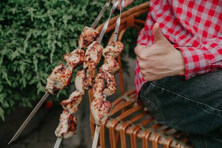 Great kebab. A man sits in a wicker chair and holds in his hand three delicious kebabs. Left hand shows a good sign.の写真素材
