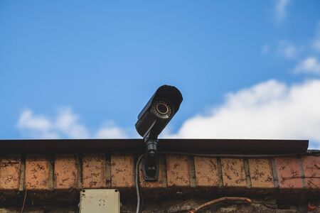 surveillance video camera is mounted on a brick fence against a blue sky with white clouds. Bottom...