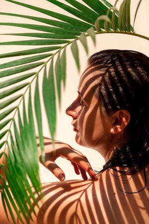 Portrait of a beautiful girl with bare shoulders without retouching with her natural imperfections. Above grows a green branch of the Howe palm tree. Palm tree gives a clear shadowの写真素材