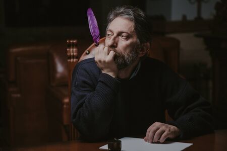 man with a beard sits at a large polished table and reflects on a white sheet. In the hand is a...