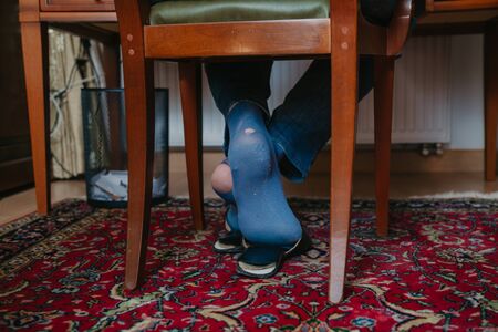 Holes in socks. A man sits on a wooden chair in the office in slippers on his both socks; you can see big holes sock, full of holes, hole, table, chair, office, carpet, furniture,の写真素材