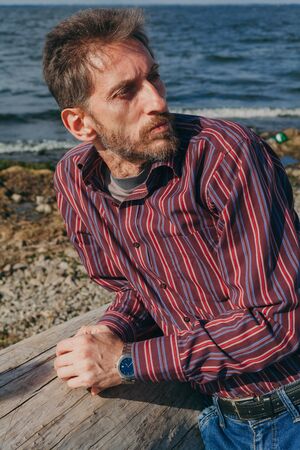 Serious adult man with a beard on the riverbank on a large old log looks thoughtfully in front of...