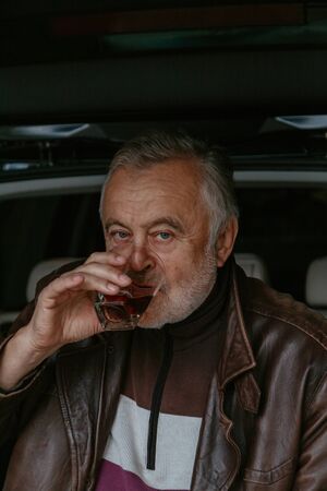 elderly man with a gray beard in a leather jacket drinks whiskey from a glass glass against the...