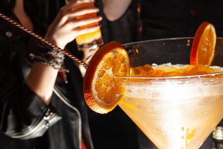 On the edge of a large, foggy martini glass, close-up round orange slices are weighed. In a glass of alcoholic cocktail with ice. Sticking out a twisted cocktail spoonの写真素材