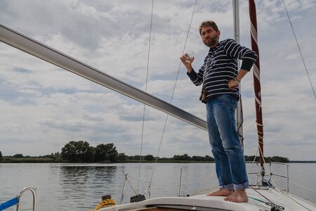 Businessman with a beard in a striped sweater stands on the nose of a white yacht and drinks whiskey from a glassの写真素材