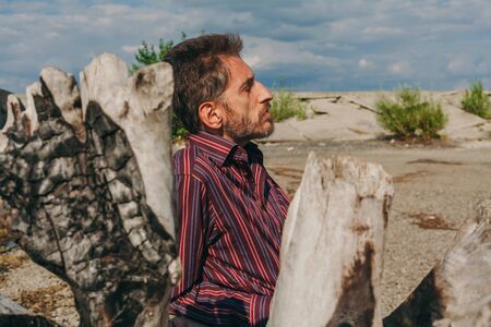 Serious adult man with a beard on the riverbank on a large old log looks thoughtfully in front of himの写真素材