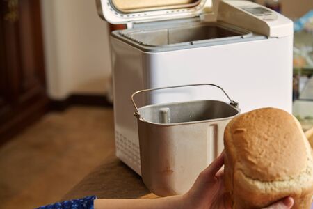 woman hands hold fresh bread from the bread baking mold of the electric bread maker. There is a big...