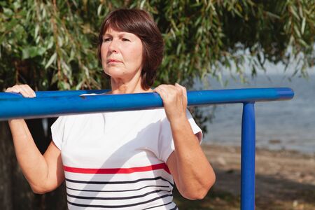 Gymnastics on the river bank. Cute elderly woman performs health exercises on metal bars