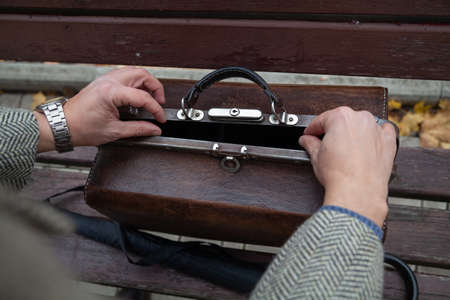 Close-up of a man in a gray coat opens a brown carpetbag on a wooden bench. View from aboveの写真素材
