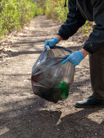 We save nature from garbage. Ecologist in blue rubber gloves collects trash in a black plastic bag in the forestの写真素材