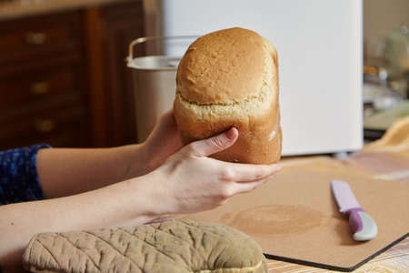 woman hands hold fresh bread from the bread baking mold of the electric bread maker. There is a big...