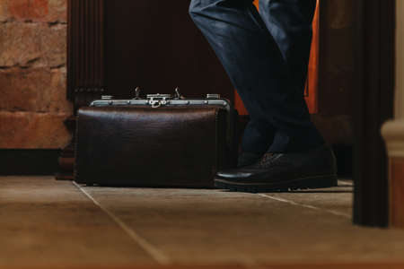 Gentleman style. A vintage travel bag stands on the floor covered with ceramic tiles. Near the legs of a man in trousers and bootsの写真素材