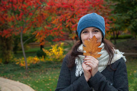 beautiful young woman in a knitted hat stands in the middle of an autumn park. Holds a yellow leaf of a tree. A female portrait without retouching with her natural imperfectionsの写真素材