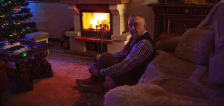 New Year Eve by the fireplace. An elderly man sits on a carpet in front of a fireplace in a dark room. A fire burns brightly in the fireplace. There is a decorated Christmas tree in the corner of the roomの写真素材