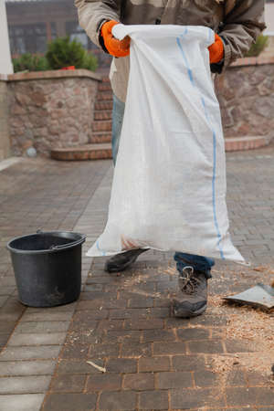 Hands in orange gloves raise a white plastic trash bag over the paving slabsの写真素材