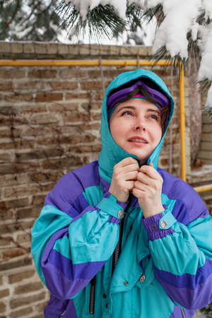 Beautiful young woman in ski overalls puts on a hood in front of a brick fence. There is snow on the...