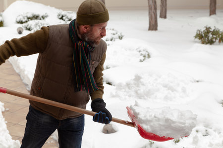 handsome man in a sports cap and a vest holds a red shovel with snow in his handの写真素材