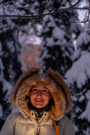 beautiful young woman in a light jacket with a hood looks into a mobile phone on the winter street....