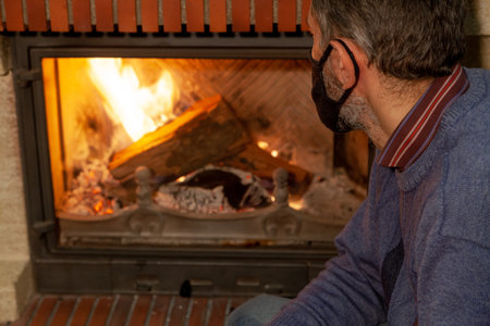 We are sitting at home on self-isolation. A man in a black protective mask looks at the fire in a burning fireplaceの写真素材