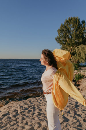 Portrait of a pretty plus size girl with long red hair and a bright yellow jacket on the sandy beach of a wide riverの写真素材
