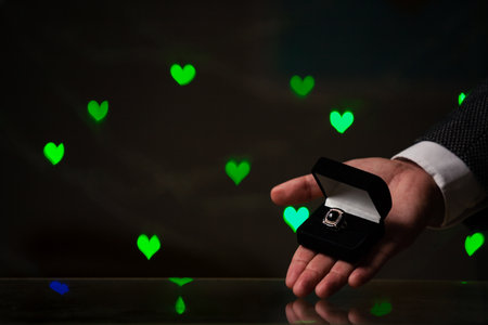 Valentine's Day gift. A man hand holds a ring in a black box. Behind on a dark background green heartsの写真素材