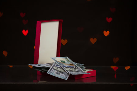 Valentine's Day gift. An open red gift box stands against a dark background. It contains a bundle of dollar bills. Multicolored hearts are glowing behindの写真素材