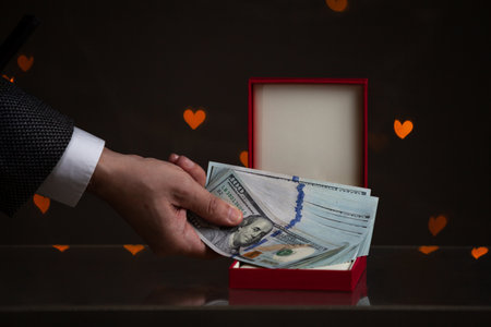 Valentine's Day gift. A hand puts a bundle of dollars in a red box. Yellow hearts are glowing behind on a dark backgroundの写真素材