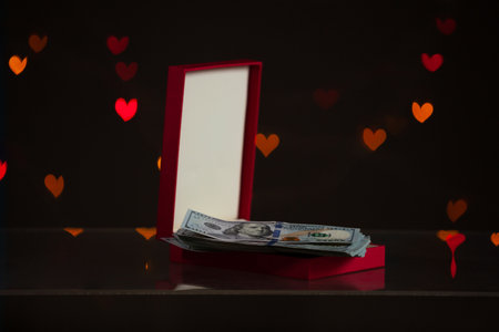 Valentine's Day gift. An open red gift box stands against a dark background. It contains a bundle of dollar bills. Multicolored hearts are glowing behindの写真素材