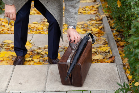 antique leather valise stands on the step in the autumn park. On top is an umbrella cane. A gentleman in a light coat touches him with his handsの写真素材