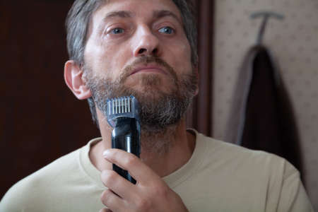 Haircut beard trimmer. A man cuts his gray beard on his own using a machine with a nozzle close-upの写真素材