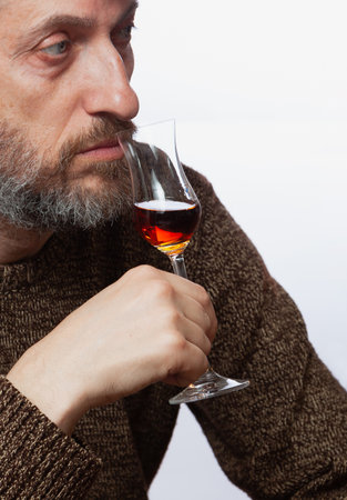 Grappa tasting. Man with gray beard in brown sweater sniffing glass goblet with grappa close upの写真素材