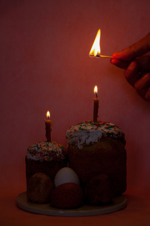 Two Easter cakes and eggs stand on a pink background. Candles are inserted into the Easter cakes. A hand lights a candle with a match. In the foreground is a tree branch with white flowersの写真素材
