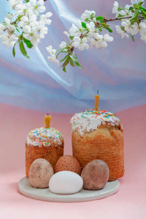 Easter still life. Two Easter cakes and eggs stand on a round stand on a pink background. Candles...