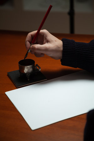Vintage writing instrument. The right hand lowers the wooden pen with the metal pen into the inkwell. Left hand holds a white sheet of paperの写真素材