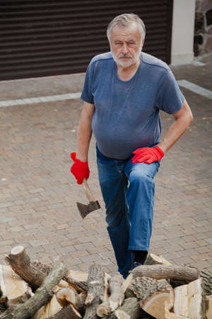 elderly man with a beard in red gloves is standing in the yard in front of a pile of firewood. Holding an axの写真素材