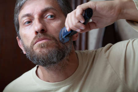 Haircut beard trimmer. A man cuts his gray beard on his own using a machine with a nozzle close-up