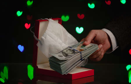 gift for Valentine's Day during the pandemic. A hand holds a bundle of dollars over a red box. Above is a medical mask. Behind on a dark background, multi-colored hearts glowの写真素材